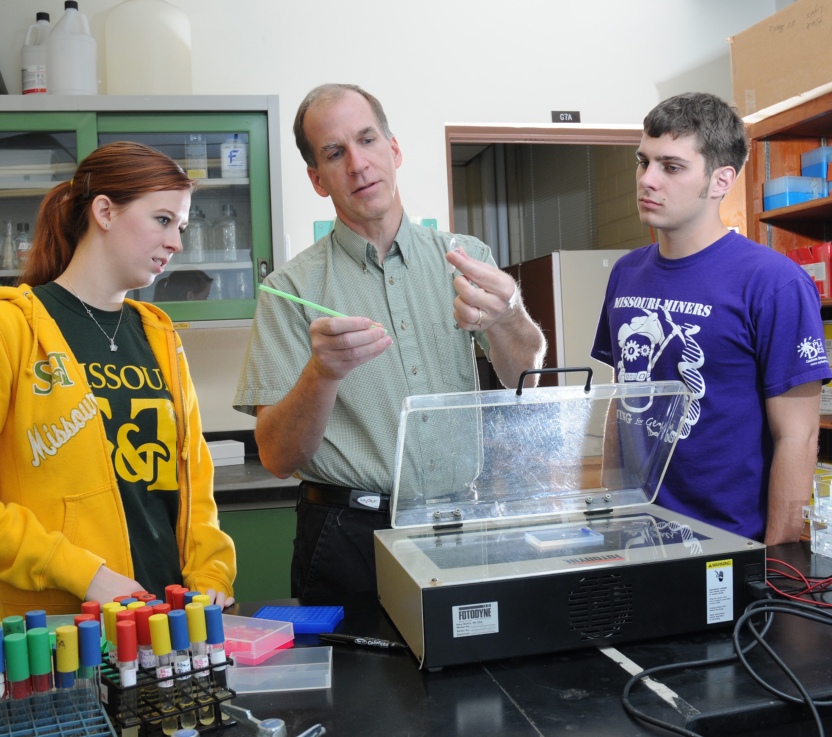 Dr. David Westenberg holding a tube and two students standing beside him, one in a yellow shirt and one in a purple shirt.